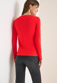 Rotes Langarmshirt mit rundem Ausschnitt, taillierter Passform und glatter Textur. Kombiniert mit dunkelgrauen Jeans mit Gesäßtaschen.