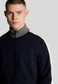 Giacca a zip blu navy con colletto grigio; presenta strisce bianche a contrasto lungo le spalle; tessuto morbido e testurizzato.