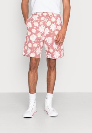 Homme portant un short fleuri rose, un t-shirt blanc, des chaussettes blanches et des baskets montantes blanches, debout les mains dans les poches.