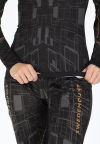 Completo aderente nero con un motivo geomtrico grigio scuro, caratterizzato da dettagli dorati. Il materiale sembra elastico, con una tasca con zip.