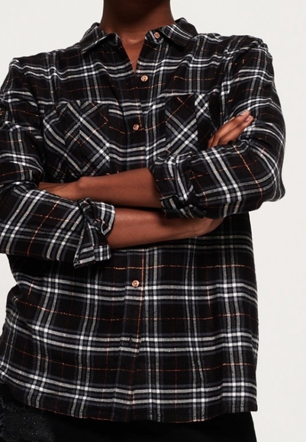 Personne à la peau foncée portant une chemise à carreaux noire, blanche et orange à boutons, manches retroussées et bras croisés.