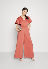 Ροζ ολόσωμο φόρεμα με φαρδιά παντελόνια και κοντά φτερωτά μανίκια, με βαθύ V ντεcolleté και ζωνάκι στη μέση, συνδυασμένο με μία σSparkly τσάντα.