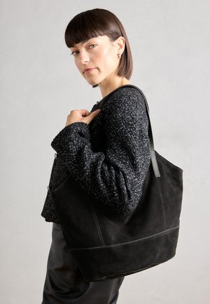 Sac fourre-tout en daim noir avec une texture lisse, une forme structurée et des coutures contrastantes. Il dispose de longues poignées pour le port à l'épaule.