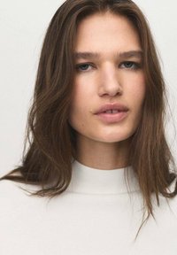 Modella con lunghi capelli castani che indossa un top bianco a collo alto. Presenta pelle liscia e trucco neutro, con uno sfondo bianco semplice.