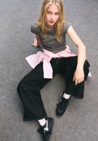 Femeie blondă așezată pe asfalt, purtând un tricou gri ajustat, pantaloni negri evazați, o cămașă roz legată în jurul taliei și adidași negri, privind în sus.