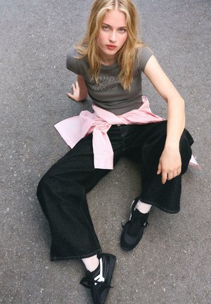 Femeie blondă așezată pe asfalt, purtând un tricou gri ajustat, pantaloni negri evazați, o cămașă roz legată în jurul taliei și adidași negri, privind în sus.
