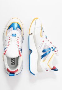 Ett par vita sneakers med blå, röda och gula accenter, meshnätpaneler och tjocka texturerade sulor, visade från ovan och sidan.