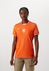 Orange fotbollsjacka med prickigt mönster, med en vit Nike-logotyp och en lejonemblem. Korta ärmar och rund krage med röda och blå detaljer.