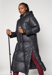 Lång svart puffjacka med huva, med dragkedja fram, sidoknappar och kviltad textur. Stylad med vandringsstavar och röda leggings.