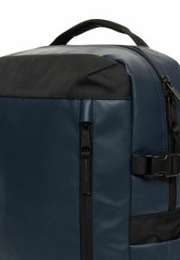 Mochila azul con un acabado liso y texturizado, parte superior de malla negra, cremallera lateral y herrajes de correas ajustables. Diseño compacto y moderno.