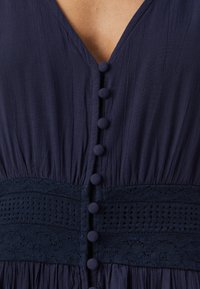 Μπλε ναυτικού φόρεμα με V λαιμόκοψη, υφή, ρέουσα ύφανση, διακοσμητικά κουμπιά μπροστά και ζώνη με δαντέλα.