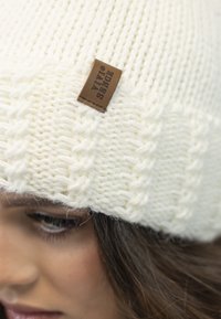 Gorro blanco de punto con un patrón texturizado y una etiqueta de cuero marrón que dice "Vivi Sence." Material suave, parecido a la lana.