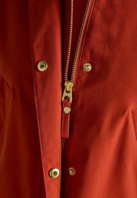 Czerwona kurtka z złotym zamkiem błyskawicznym i zatrzaskami, wykonana z gładkiego materiału o lekko fakturowanym wykończeniu. Zbliżenie ukazujące szczegóły.