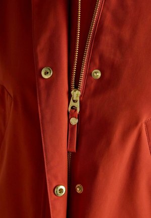 Rote Jacke mit goldenem Reißverschluss und Druckknöpfen, aus glattem Stoff mit leicht strukturierter Oberfläche. Nahaufnahme, die die Details hervorhebt.