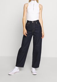 White sleeveless top with high collar and front zipper paired with dark blue wide-leg jeans and white sneakers. Minimalistic design and classic fit.