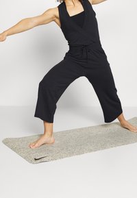 Combinaison de sport noire avec un design portefeuille, présentant des jambes raccourcies. Le modèle se tient sur un tapis de yoga gris moucheté. Position pieds nus.