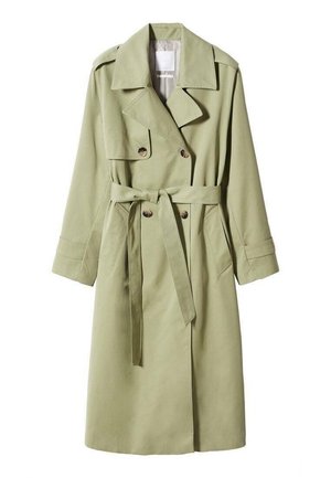 Personne portant un pull vert zippé, un trench-coat olive, un jean bleu et des bottes noires à bout pointu debout sur un sol clair.