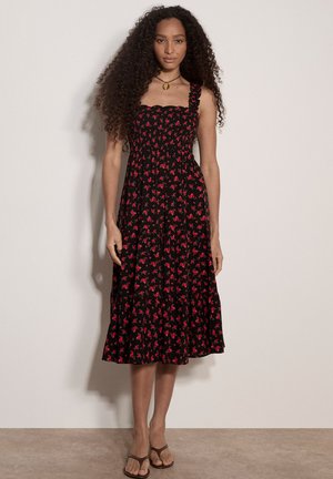 Mujer con cabello largo y rizado que lleva un vestido midi negro con estampado floral rojo y sandalias marrones, de pie contra una pared lisa.