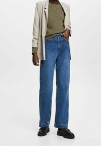 Blazer gris clair sur un pull texturé vert olive, associé à un jean en denim bleu taille haute et des chaussures noires à enfiler.