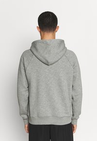 Homme portant un sweat à capuche gris uni, debout dos à l'appareil photo sur un fond neutre.