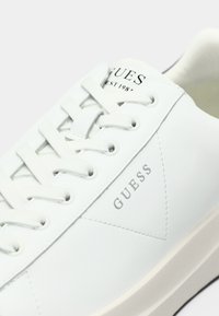 Sneaker in pelle sintetica bianca con punta rotonda, suola in gomma spessa e lacci piatti. Presenta il logo "GUESS" in rilievo e un dettaglio di design triangolare.
