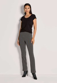 Vrouw staat tegen een bleke achtergrond, gekleed in een zwart T-shirt met korte mouwen, zwarte en witte chevronpatroon broek, zwarte hoge laarzen en gouden oorbellen.