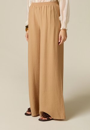 Beige bukser med vidde ben og elastisk talje, båret med en hvid bluse med lange ærmer og sorte flade sandaler med stofsløjfer.