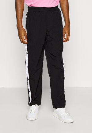 Persoon die zwarte sportbroek draagt met witte drukknopstrepen aan de zijkanten en witte sneakers, staand tegen een effen achtergrond.