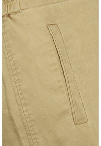 Gros plan sur un tissu en lin beige présentant une poche latérale aux bords cousus, affichant une texture lisse et de légères rides.