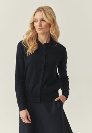 Jeune femme aux longs cheveux blonds portant un cardigan noir boutonné et une jupe, debout avec une main dans la poche sur un fond neutre.