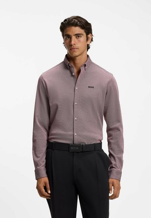 Joven con una camisa ajustada de manga larga color malva abotonada y pantalones negros con cinturón negro sobre un fondo blanco liso.