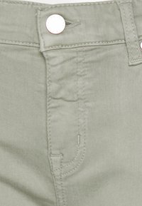 Jeans de algodón verde con una textura lisa, que cuentan con bolsillos frontales, un cierre de botón metálico y sutilezas en los detalles de costura.