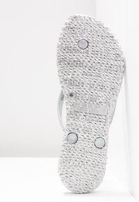 Silverfärgad toffel med strukturerad sula, prydd med präglade varumärkesdetaljer och två runda metallackenter. Lätt och flexibel design.