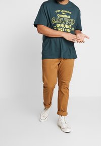 Donkerblauw-groene katoenen T-shirt met gele grafische tekst, gecombineerd met opgerolde kaki-broeken en witte hoge sneakers op een lichte achtergrond.