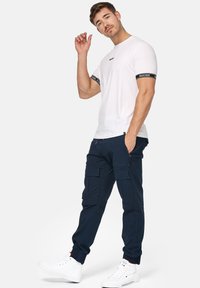 Camisa blanca de manga corta con acentos de logo en blanco y negro; pantalones cargo azul marino con bolsillos; zapatillas deportivas altas blancas.