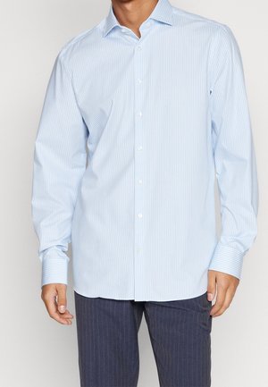 Mann trägt hellblau vertikal gestreiftes Hemd und dunkle Nadelstreifenhose, steht vor schlichten Hintergrund.