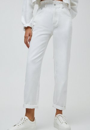Femme portant un jean blanc taille haute avec les ourlets retroussés, associé à des baskets blanches et un sweat-shirt blanc court.