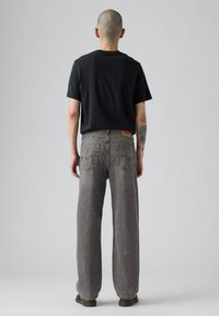 Sive široke traperice ravnog kroja s opuštenim fitom, s kožnim patentnim dijelom na struku i stražnjim džepovima s ukrasnim šavovima, u kombinaciji s crnom majicom.