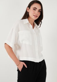 White cropped shirt in lightweight fabric, featuring a button front, short sleeves, and two chest pockets. Smooth texture.