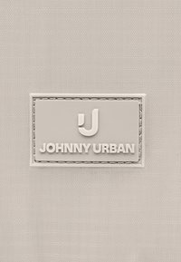 Etichetta in tessuto beige con bordi cuciti, presenta un logo in rilievo "J" sopra il testo "JOHNNY URBAN" in lettere maiuscole.