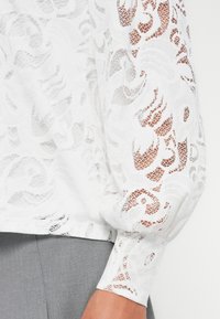 Blusa bianca in pizzo con intricate lavorazioni a giorno, maniche lunghe gonfie e polsini aderenti. Tessuto morbido, materiale leggero e design trasparente.