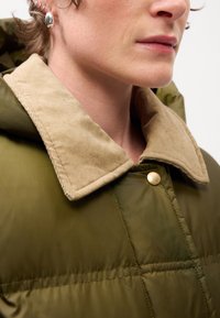 Jachetă puffer verde măsliniu cu guler din corduroy bej și detalii cu nasturi aurii, având o textură fină și un design matlasat.