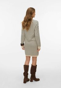 Vestito beige al ginocchio con strisce a chevron nere, maniche lunghe e polsini a costine. La modella indossa stivali marroni sopra il ginocchio.