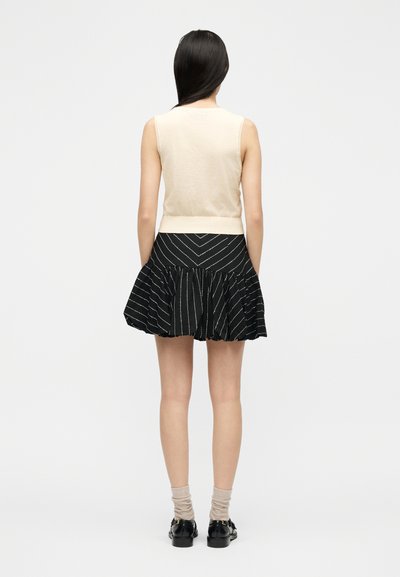 Γυναίκα με μακριά σκούρα μαλλιά φοράει αμάνικη μπεζ μπλούζα, μαύρη φουστα με άσπρες ρίγες, μπεζ κάλτσες και μαύρα μοκασίνια, με την πλάτη προς τα εμάς.