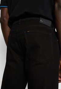 Isiku tagantvaade, kes kannab musti Lee teksaseid ja musta särki, millel on sinine triip varrukal, keskendudes teksade tagataskule ja etiketti.