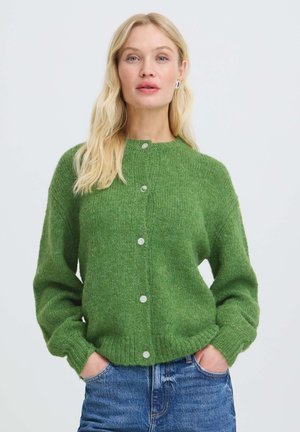 Blondine vrouw met een groene, aangeknoopte cardigan, handen in de zakken en blauwe spijkerbroek tegen een effen lichte achtergrond.
