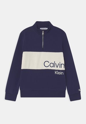 Sweat-shirt bleu marine à demi-fermeture éclair avec une bande horizontale blanche traversant la poitrine, affichant partiellement le logo "Calvin Klein" en grandes lettres.