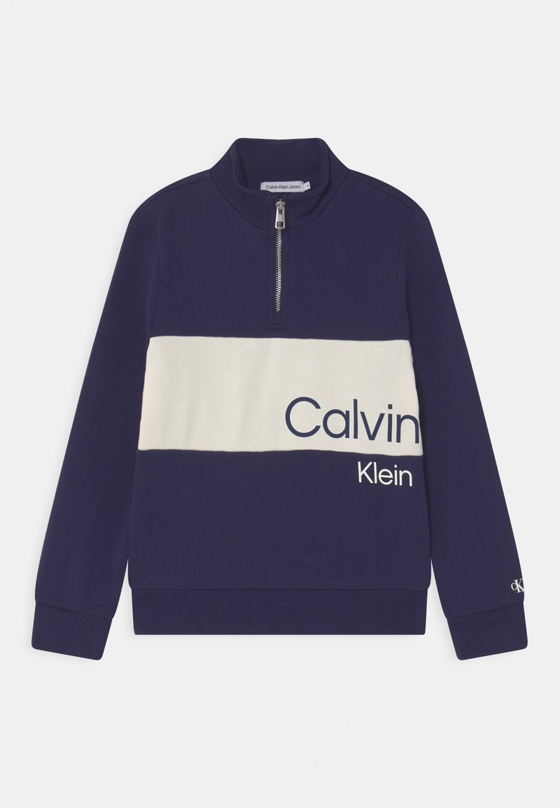 Sweat-shirt bleu marine à demi-fermeture éclair avec une bande horizontale blanche traversant la poitrine, affichant partiellement le logo "Calvin Klein" en grandes lettres.