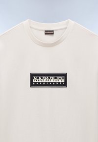 Cremefarvet t-shirt med rund halsudskæring og korte ærmer, prydet med et sort rektangulært logo med hvid skrift centreret på brystet.
