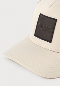 Beežikas müts struktuurse disainiga, millel on tume ruudukujuline plaastriga, millel on mustalt kujutatud sõna „BOSS" ja kergelt kaarduv nokk.